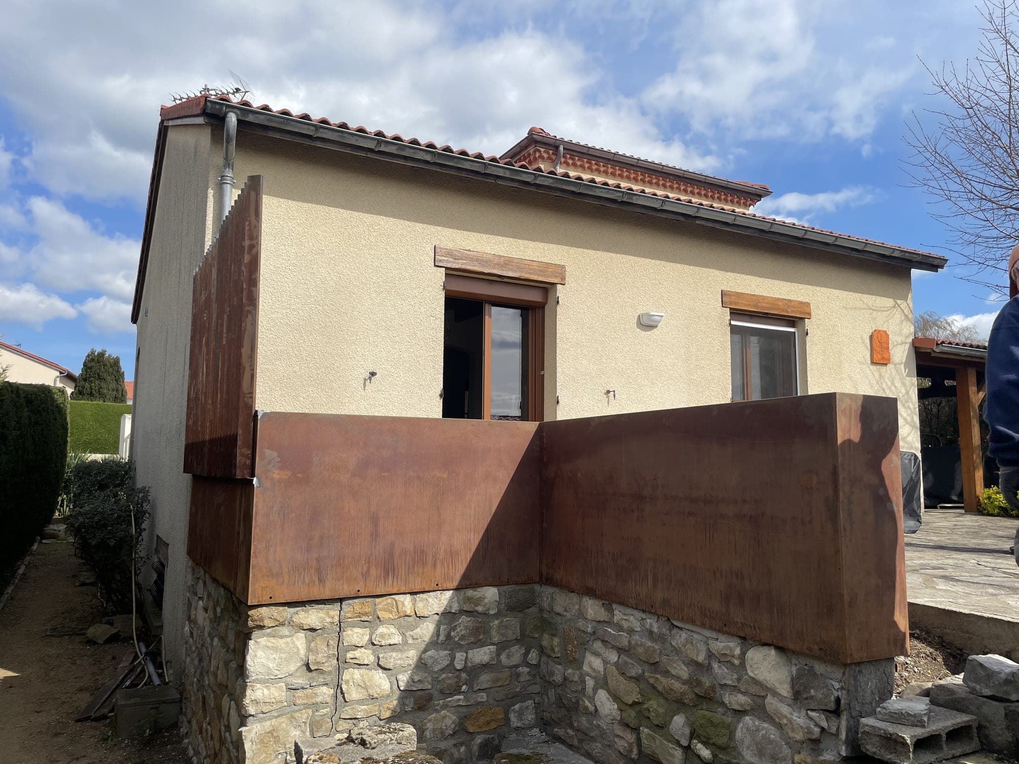 Vue extérieure : le bac en Corten borde la terrasse surélevée, au-dessus du mur de pierre