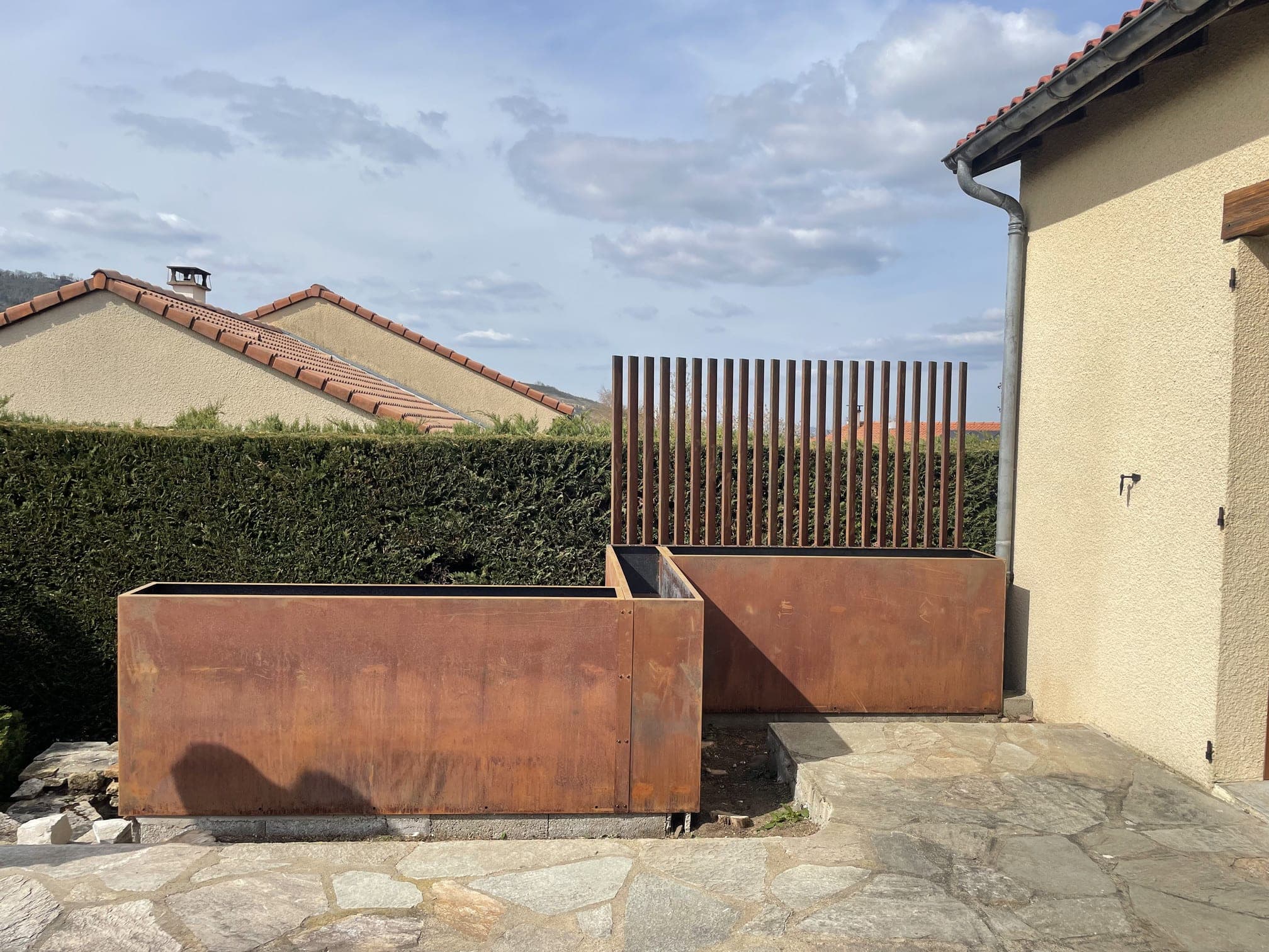 Vue frontale du bac à fleurs en acier Corten et de son claustra, devant la haie