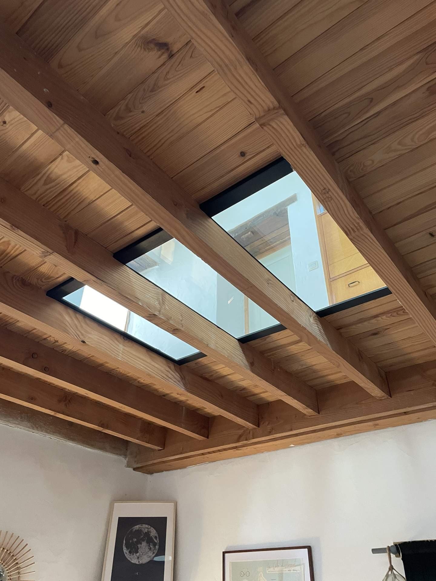 Vue du dessous : le vitrage apparaît entre les solives du plafond en bois et laisse passer la lumière