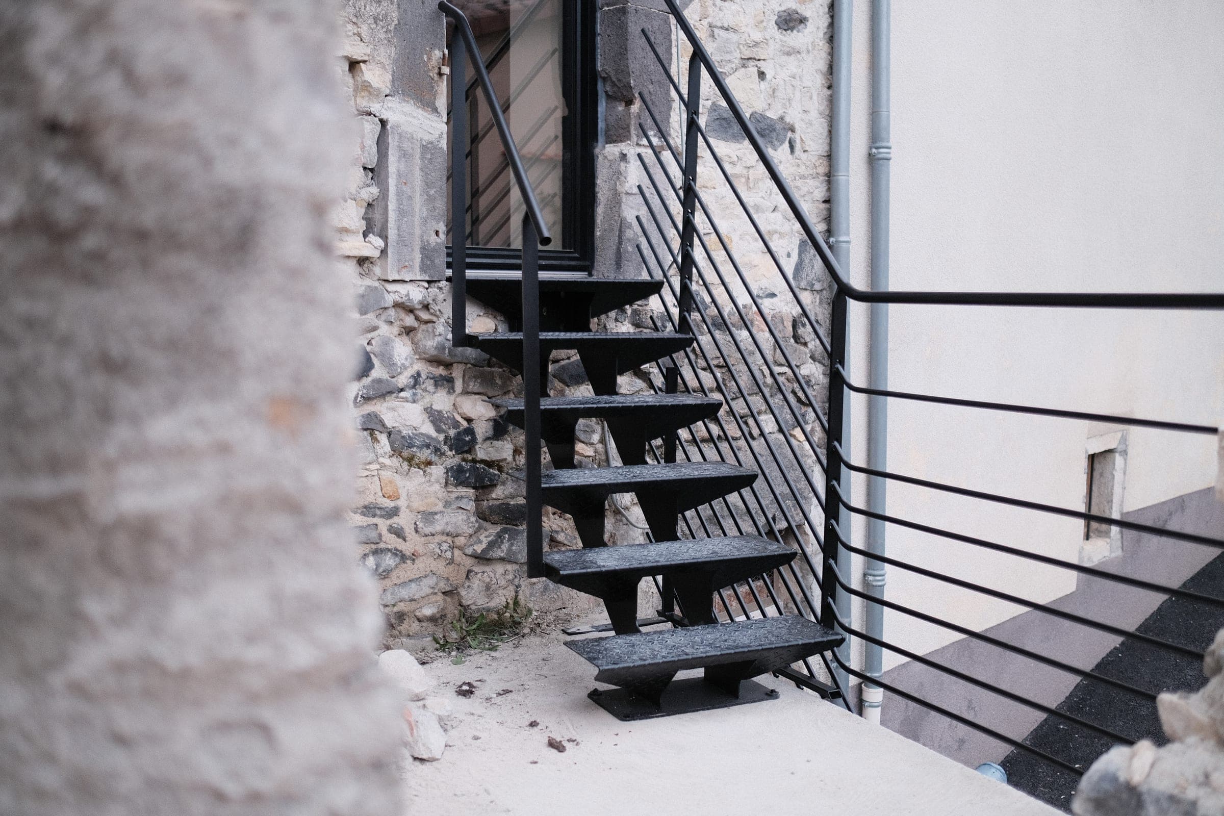 Escalier extérieur droit en acier noir à limon central, devant une façade en pierre, avec son garde-corps.