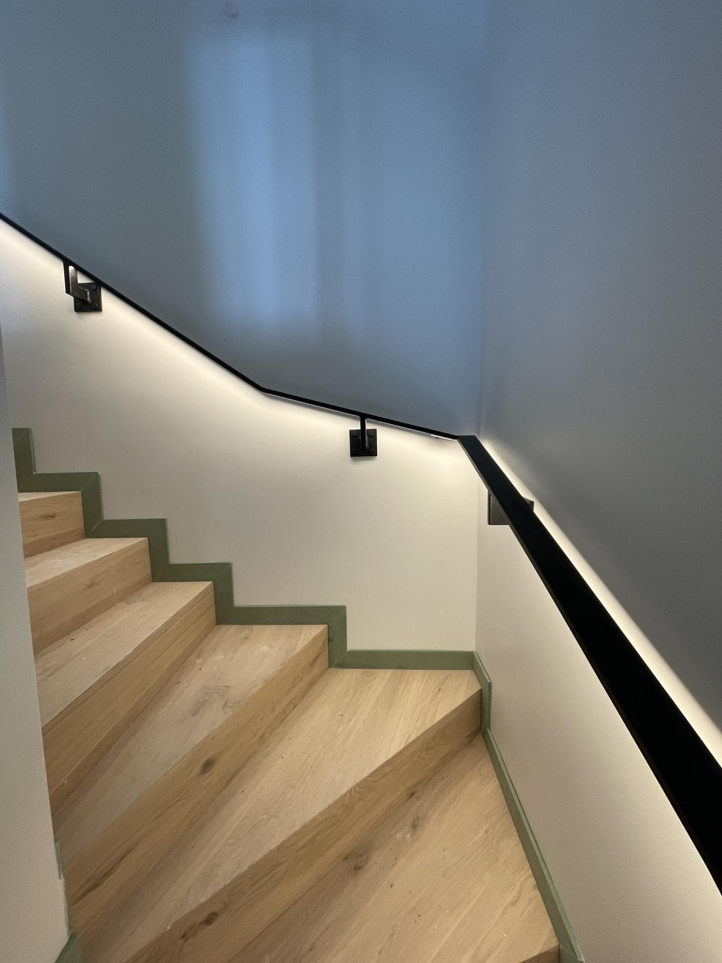Main courante en acier noir suivant la pente d’un escalier en bois clair, halo LED continu sur le mur