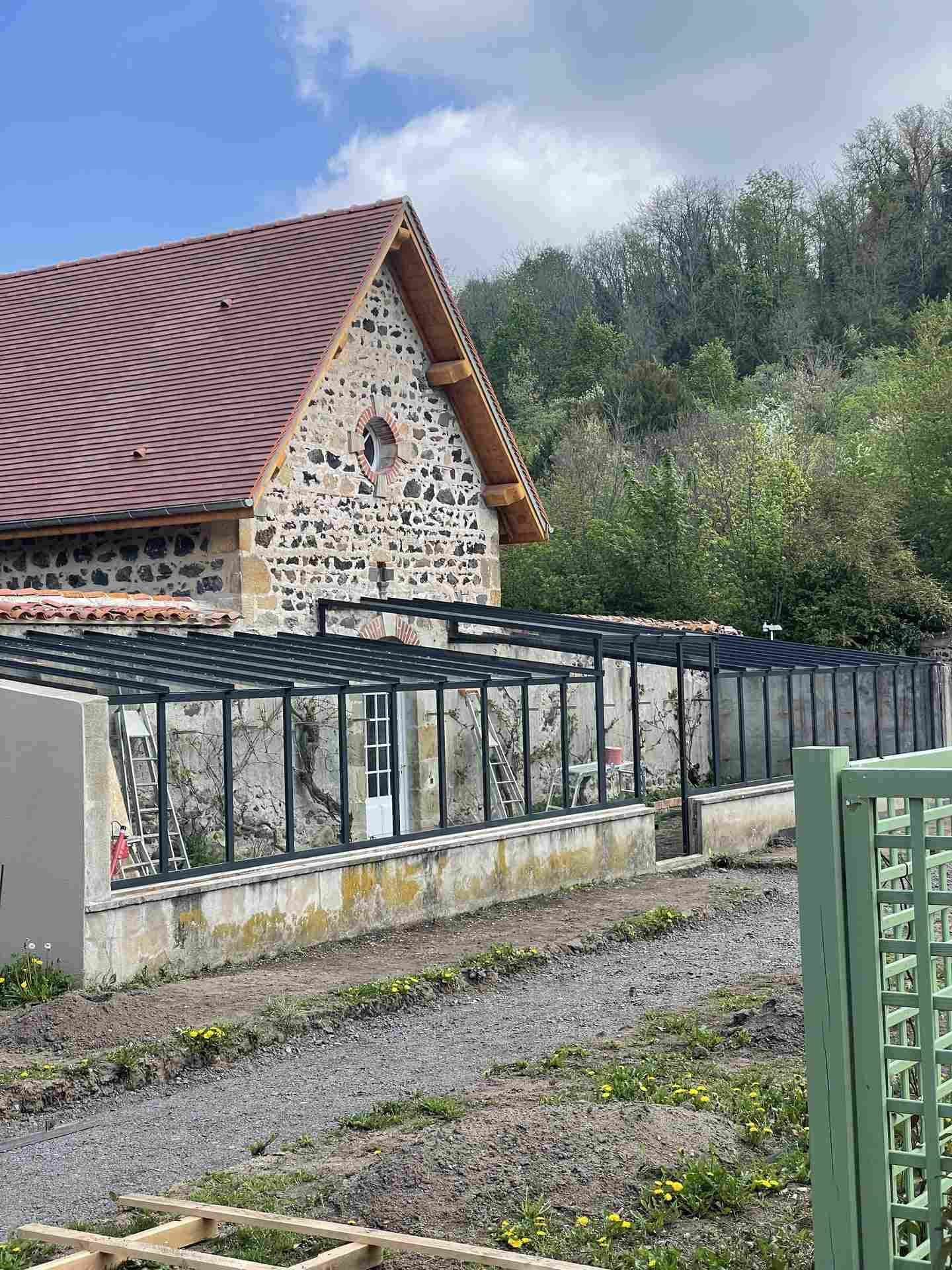 La serre vue depuis l’allée, structure gris foncé sur soubassement maçonné, bâtiment de pierre en arrière-plan