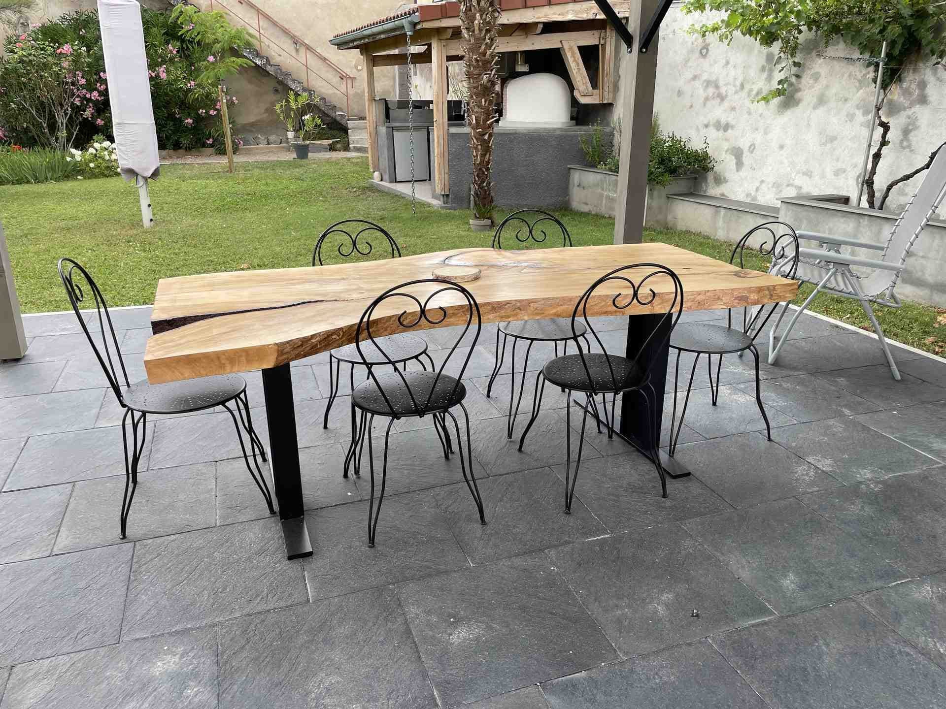 Table en tilleul massif vue à hauteur d’assise, deux pieds plats en acier noir sur semelles, jardin derrière