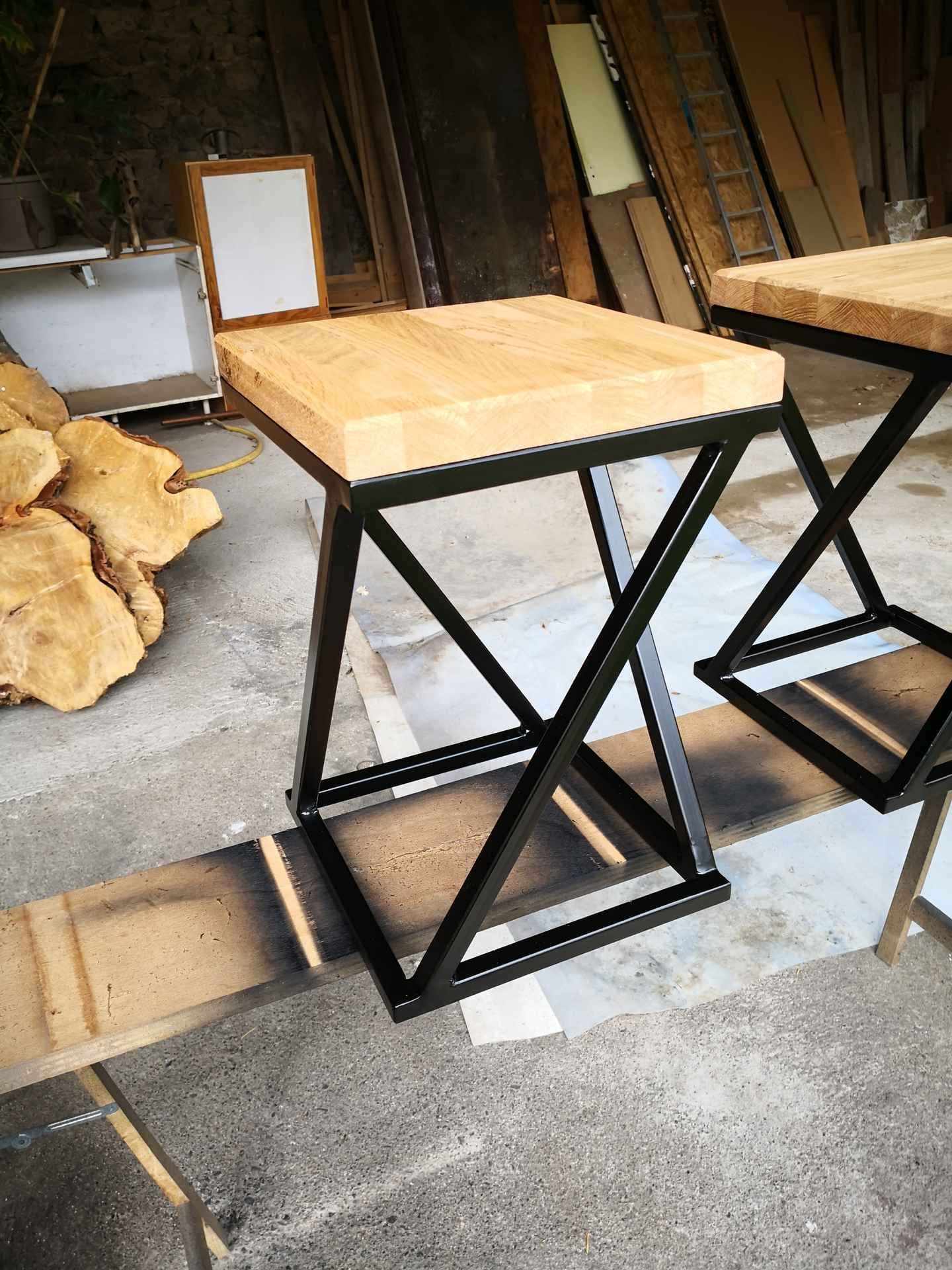 Tabouret à piétement en X, acier noir et assise en bois clair, posé sur un banc dans l’atelier