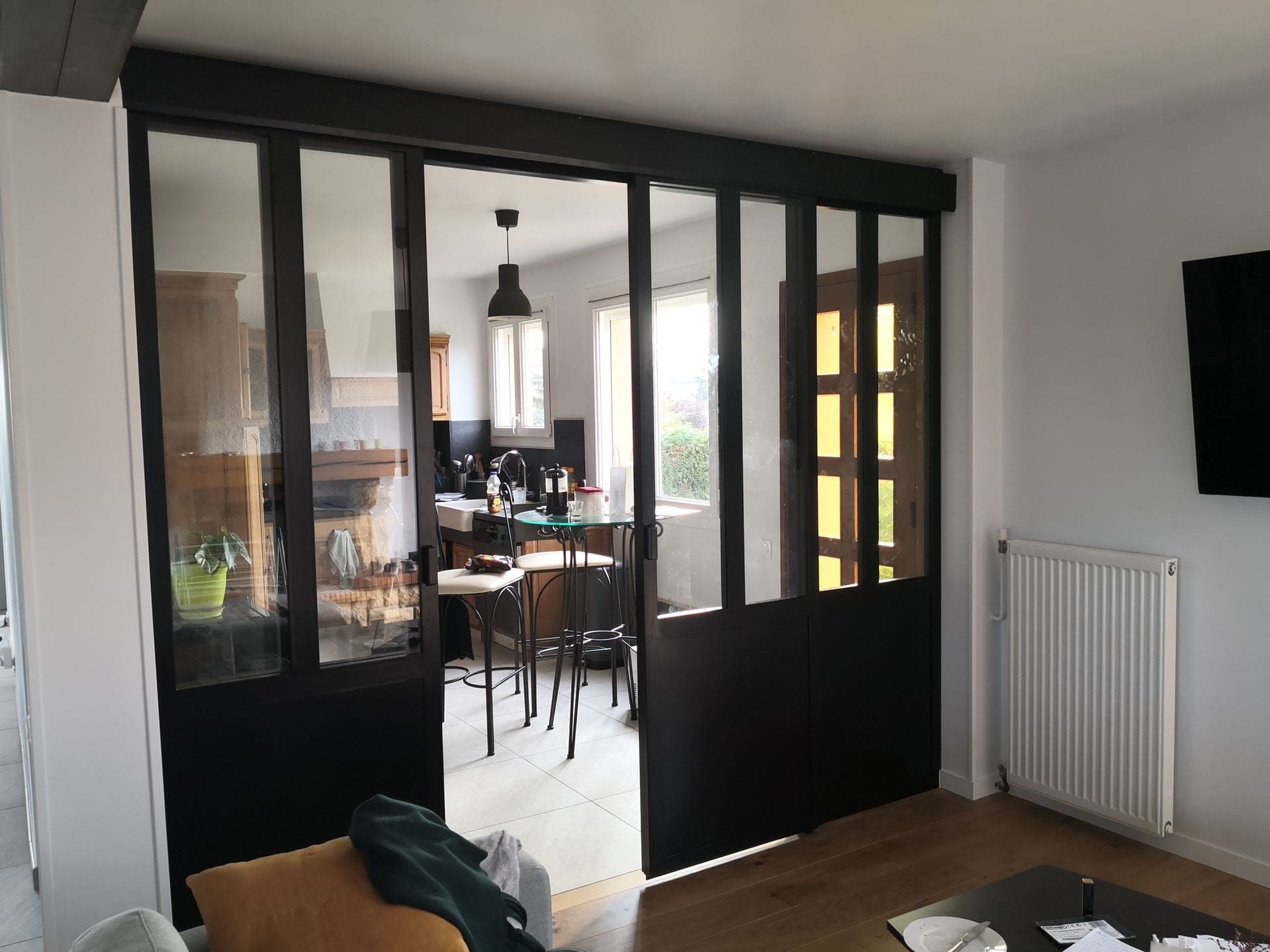 Verrière vue de biais depuis le salon, vantaux ouverts, cuisine visible à travers le vitrage