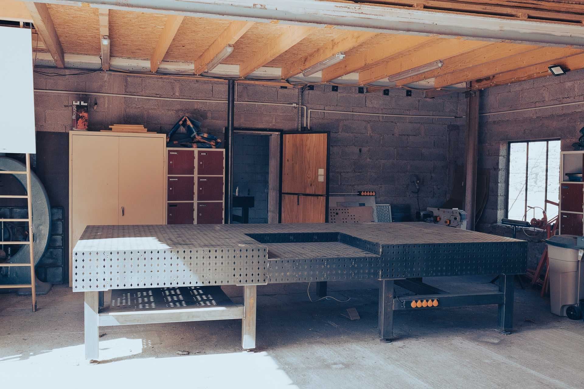 Grande table de soudure modulaire perforée dans le hall de l’atelier, sous la charpente bois.