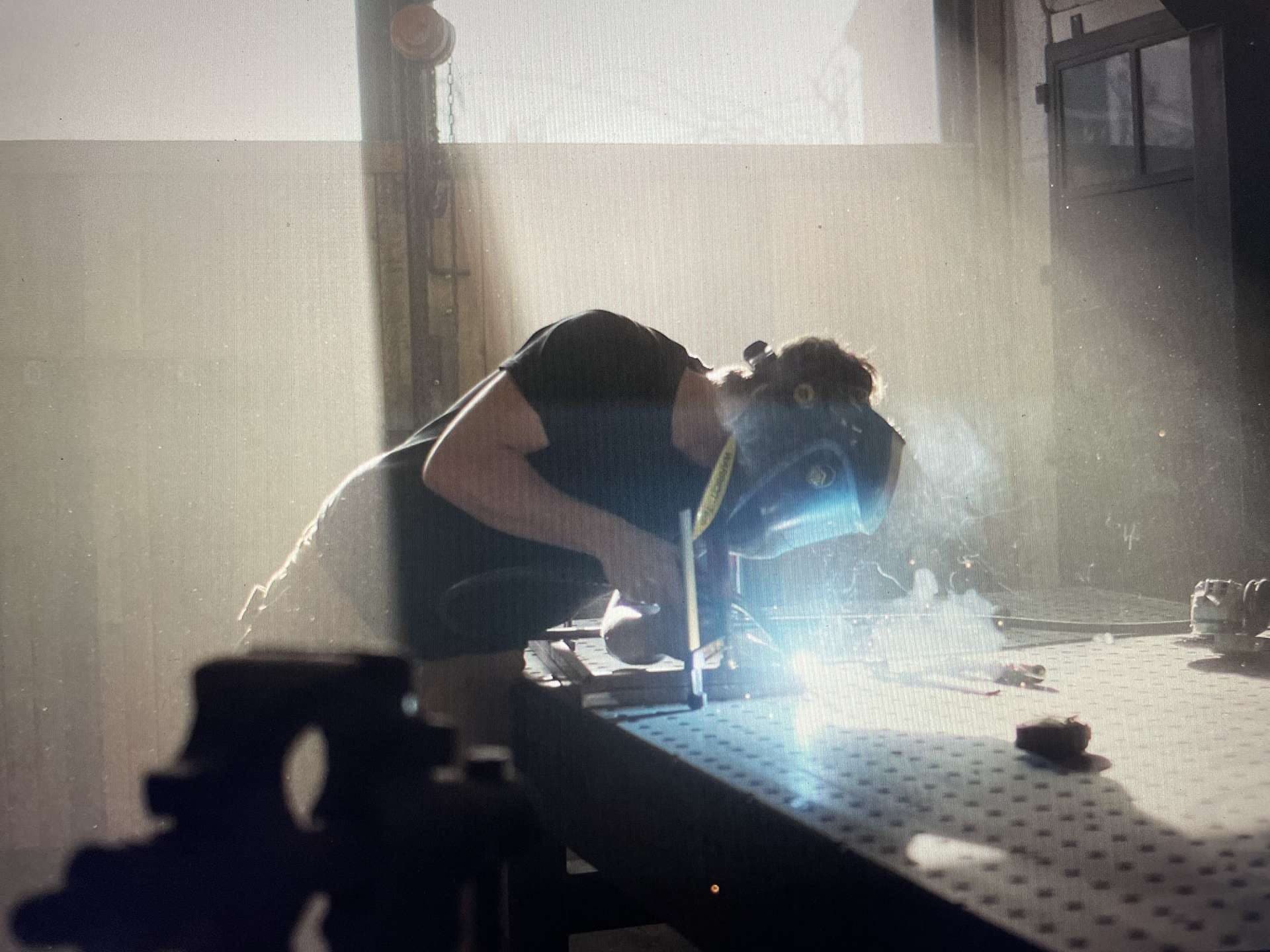 Maxence accroupi sur la table de soudure, en train de souder, éclairé à contre-jour.